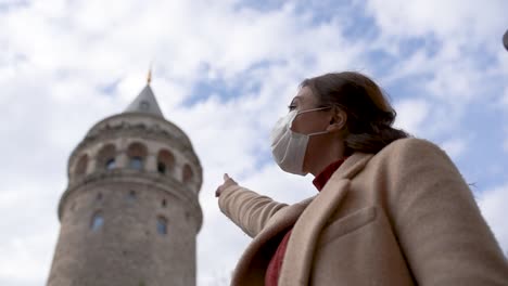 Cámara-Lenta:-Hermosa-Chica-Con-Máscara-Médica-Protectora-Y-Ropa-De-Moda-Muestra-Signos-De-La-Torre-De-Galata-Nuevo-Estilo-De-Vida-Normal,-Concepto-De-Viaje