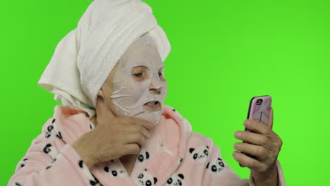 abuela en bata de baño, máscara facial. anciana usando teléfono móvil para llamada de video