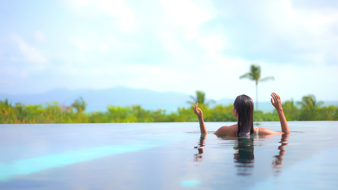 Back to the camera, a fit woman slowly rises out of the water of an infinity-edge pool while slicking back her wet hair