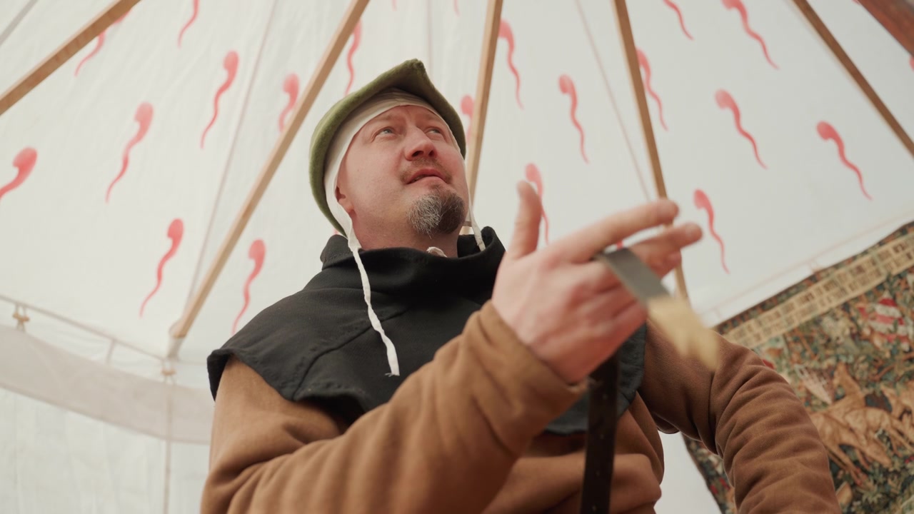 Mężczyzna w średnim wieku w średniowiecznym stroju trzyma drewnianą laskę i mówi wewnątrz namiotu z wzorzystego płótna, z ekspresyjnym ruchem ręki, miękkim światłem dziennym i nastrojem szczerego historycznego przedstawienia