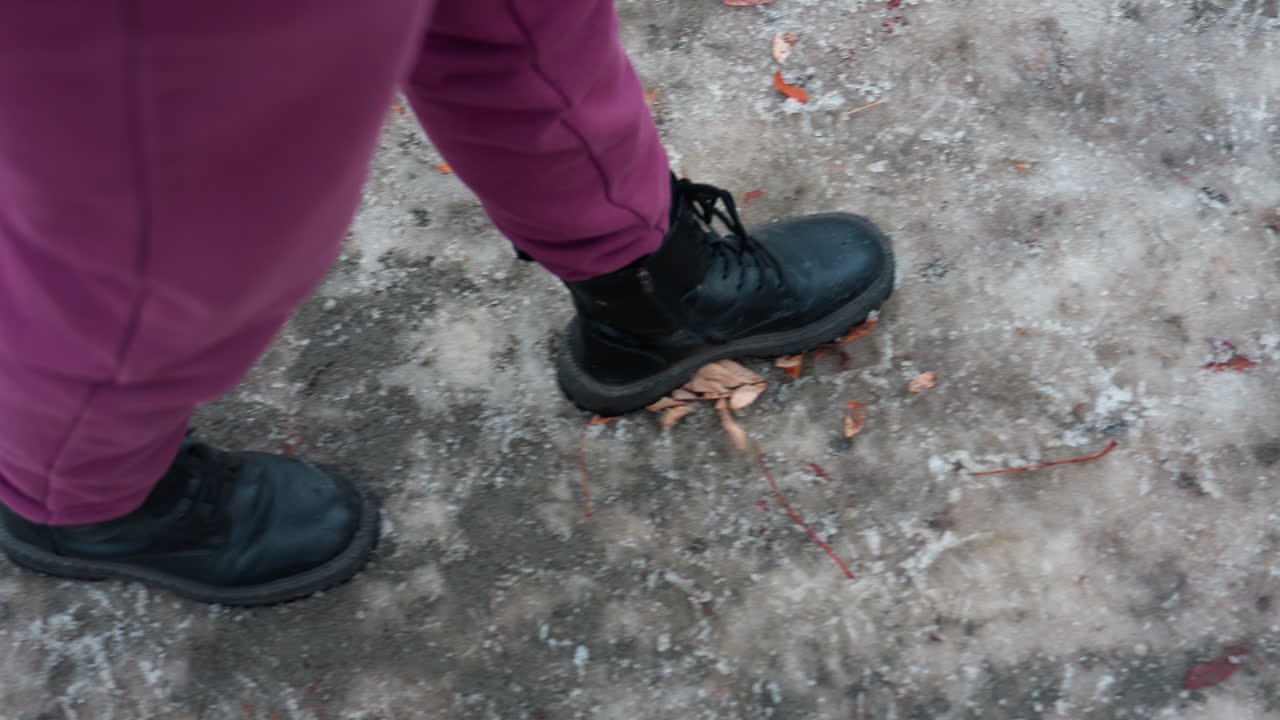 primer plano de la parte inferior de las piernas con pantalones marrones y botas negras caminando por un camino helado y desgastado cubierto de follaje esparcido, huellas impresas en el suelo congelado