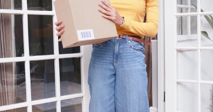 Donna con un top giallo che nota una scatola di cartone con codice a barre sulla soglia di casa, la raccoglie e la porta dentro.
