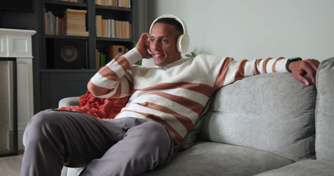 Uomo afroamericano che inizia a controllare il telefono su un divano grigio a casa, regolando le cuffie, dando un'occhiata a sinistra.