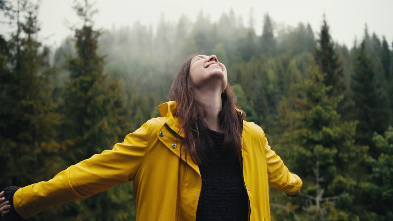 A happy girl in a yellow jacket stands against the background of a coniferous mountain forest, looks at the sky and rejoices in a little rain