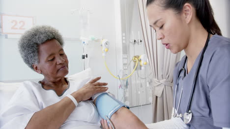 diverse female doctor testing blood pressure of senior female patient in hospital room, slow motion