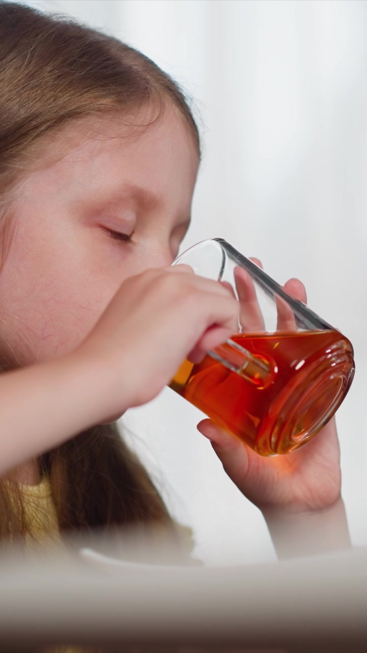 Primo piano di una bambina che beve tè da un bicchiere. La bambina tiene una tazza trasparente con liquido ambrato e sorseggia ripetutamente mentre è seduta a tavola. L'illuminazione soffusa evidenzia il suo profilo e la tazza trasparente.