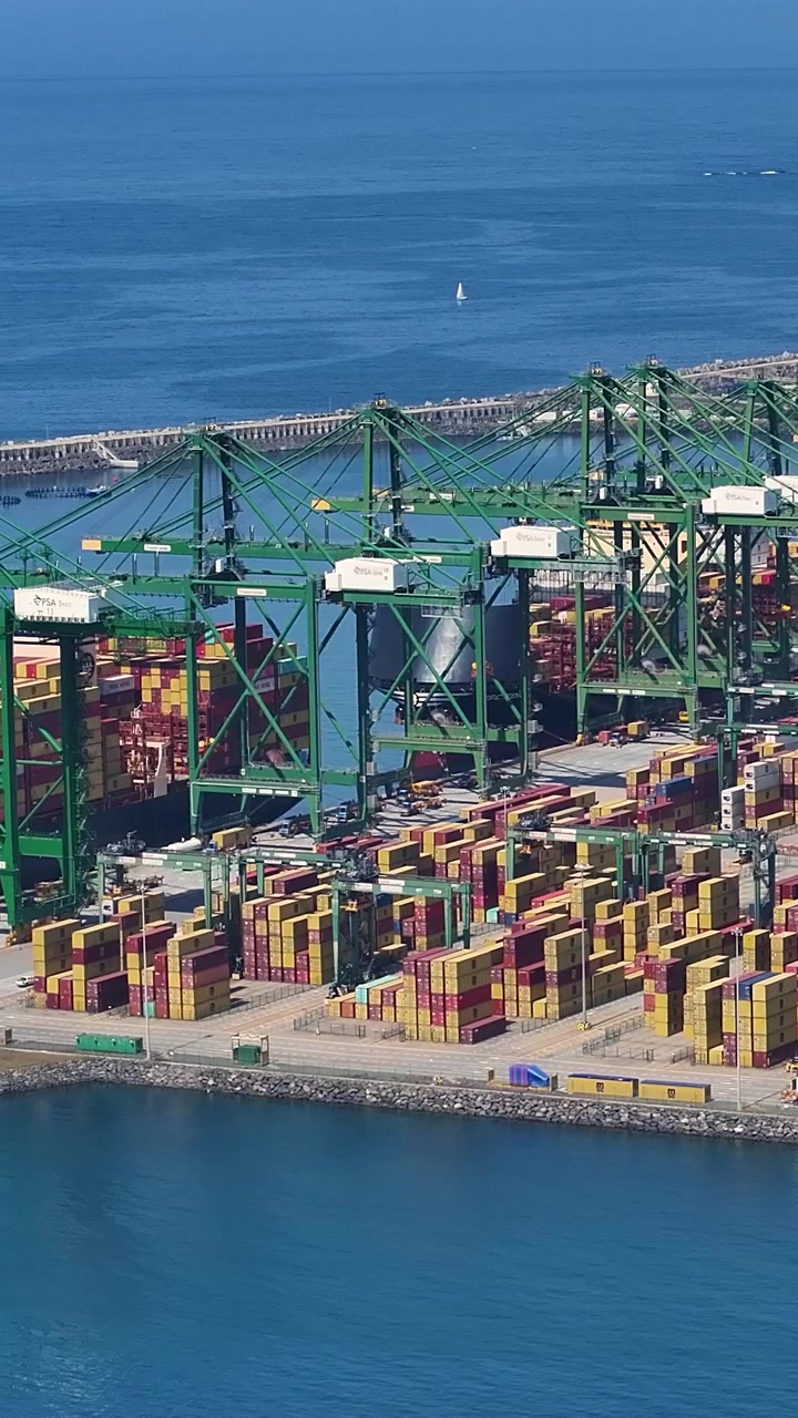 Container terminal shows shipping containers stacked for loading and unloading. Cargo ships dock along the shore at a busy port facility during daytime activities.