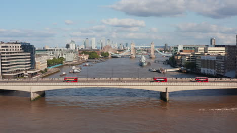 Forwards-fly-above-water-surface-of-Thames-river.-Three-red-double-deckers-driving-on-London-Bridge.-Historic-well-known-Tower-Bridge-in-background.-London,-UK