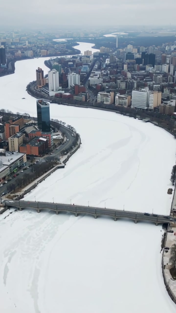 Vertical video: Panning drone surveying frozen river at city waterfront, showing bridge, ice tracks