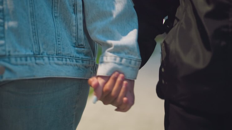 Close-up of two people holding hands outdoors, one wearing denim and the other black jacket, symbolizing connection, support, care, and emotional bond during walk in peaceful urban environment