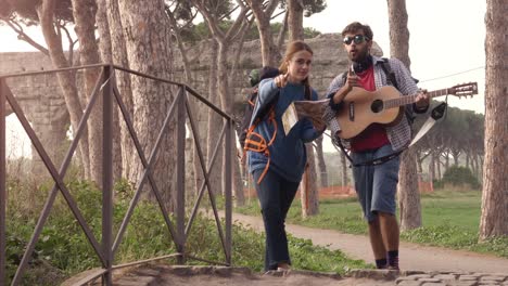 happy young couple backpackers tourists reading map guide for directions lost near roman aqueduct arches in parco degli acquedotti park ruins in rome at sunrise with guitar and sleeping bag slow motion steadycam