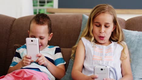 Smiling-boy-and-girl-sitting-on-sofa-and-using-mobile-phone