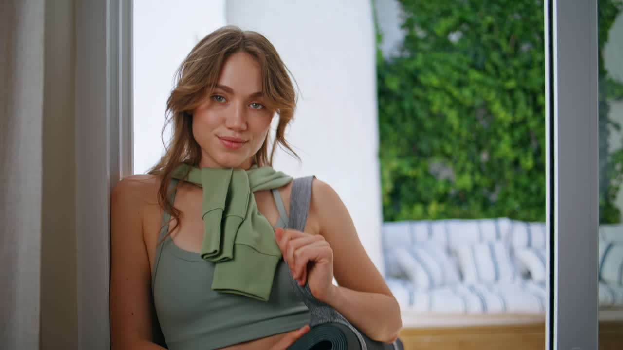 Sporty lady looking camera holding yoga mat near window closeup. Confident woman