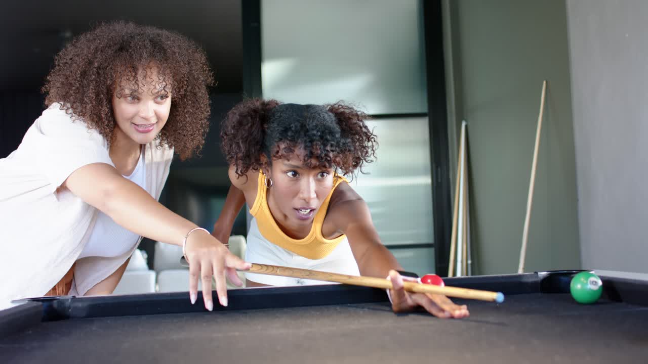 African American women at pool table, friend guiding shot, player in yellow hitting cueball for fun
