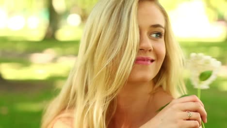 pretty blonde relaxing in the park smiling at the camera while smelling a flower