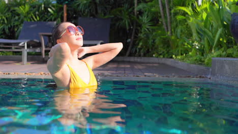 A-passionate-sensual-young-Asian-woman-touches-her-wet-hair-in-the-swimming-pool-of-an-exotic-tropical-hotel-at-sunny-day