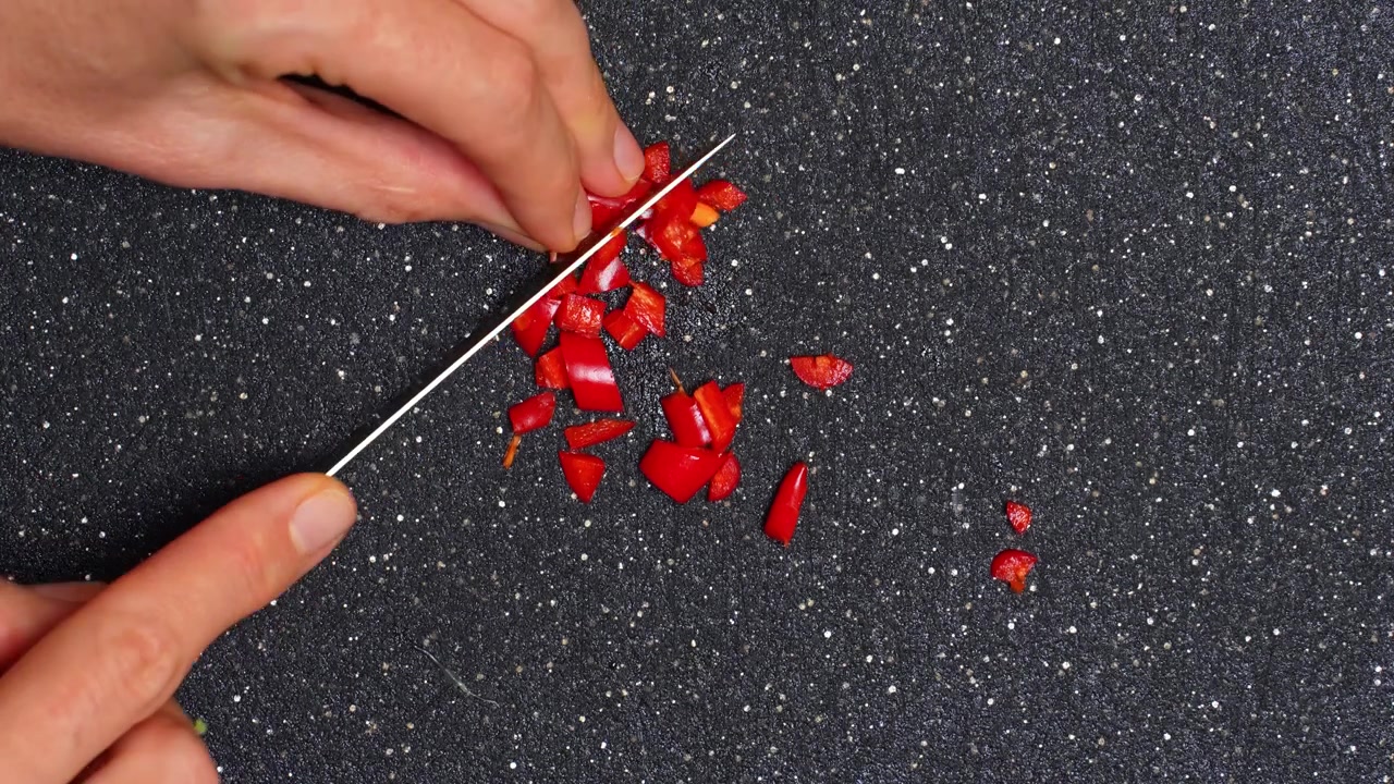Tagliare peperoni rossi su un tagliere nero in cucina. Una persona taglia i peperoni rossi a pezzetti usando un coltello affilato su un tagliere nero.