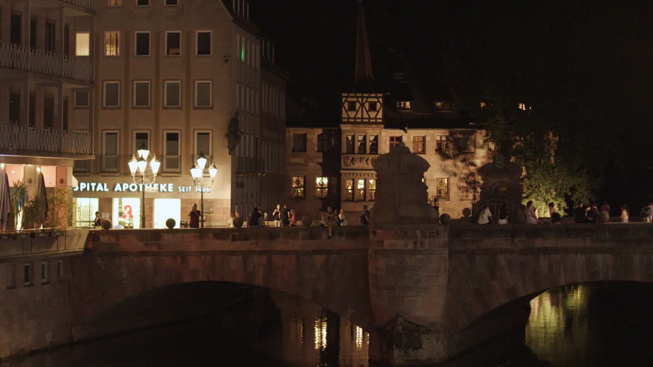 People walk across illuminated old town bridge near restaurants, warm lighting, steady wide shot