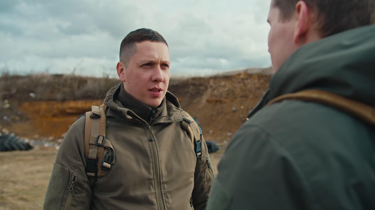 Over his shoulder a confident military man communicates with his colleague in a dark green army uniform and a backpack on his shoulders on the territory of a military training ground