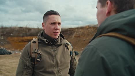 over his shoulder a confident military man communicates with his colleague in a dark green army uniform and a backpack on his shoulders on the territory of a military training ground