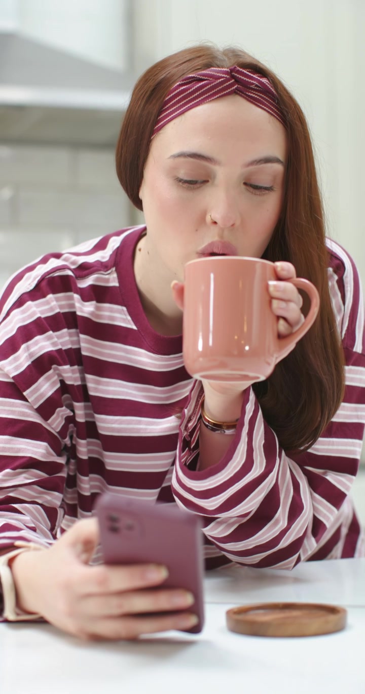 Video verticale: Donna che tiene uno smartphone color malva, legge lo schermo e poi sorseggia da una tazza color pesca al bancone