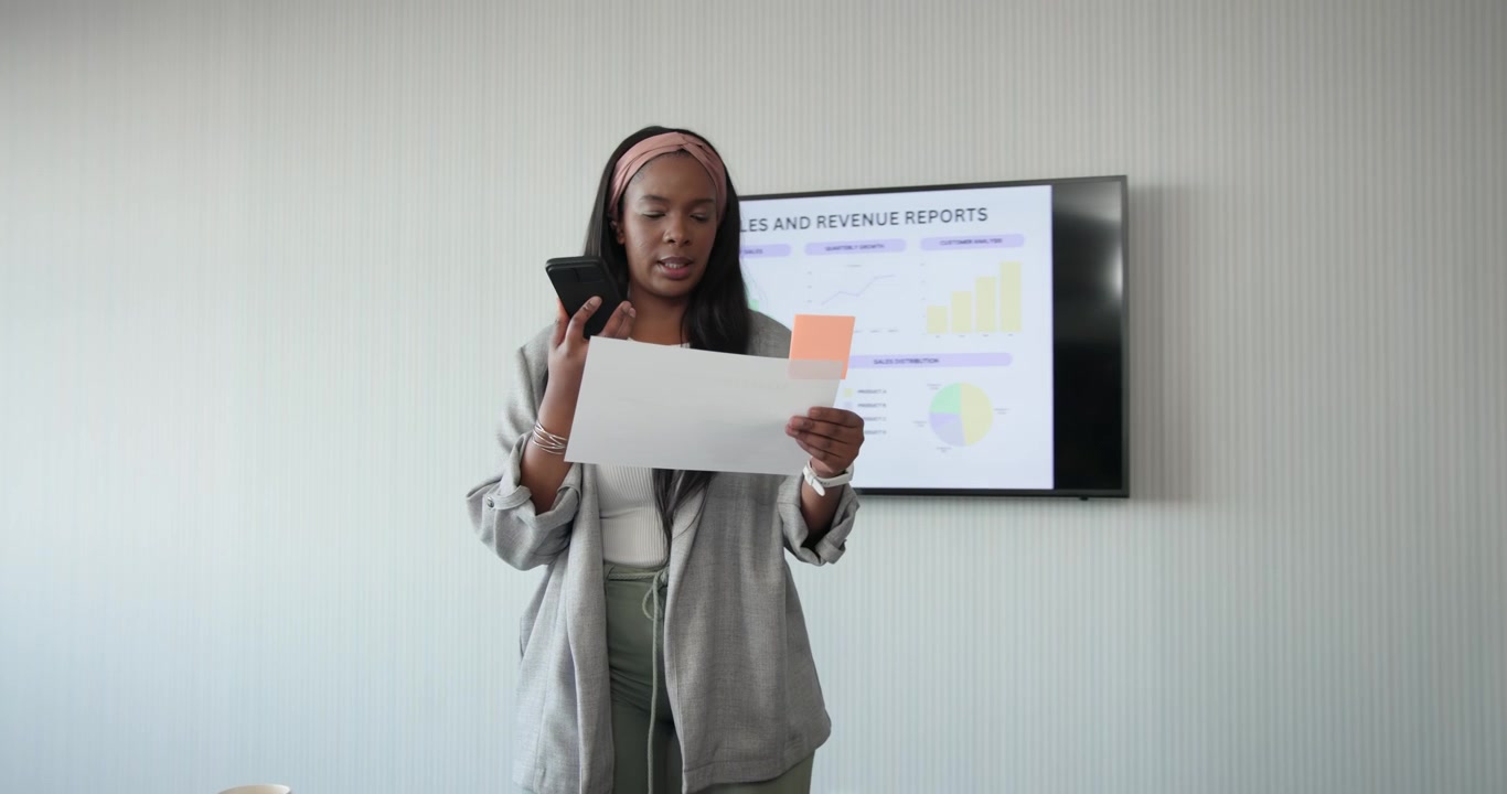 Adult African woman in blazer at table presenting Sales and Rev slide reading notes speaking into mi