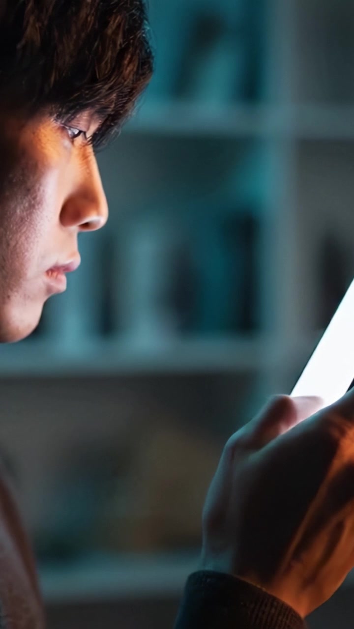Uchvácen obrazovkou: Mladý muž pohroužený do digitálního zařízení, osvětlený jeho světlem v tlumeně osvětlené místnosti s policemi v pozadí