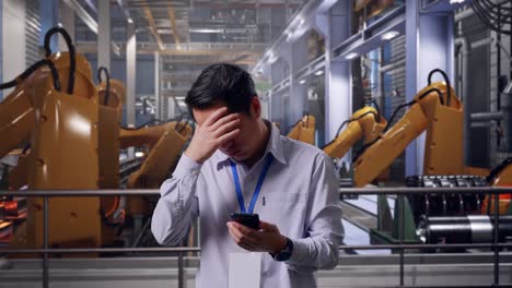 asian male professional worker standing with his smartphone with industrial robotic arms working, he is nodding his head with dissapionted