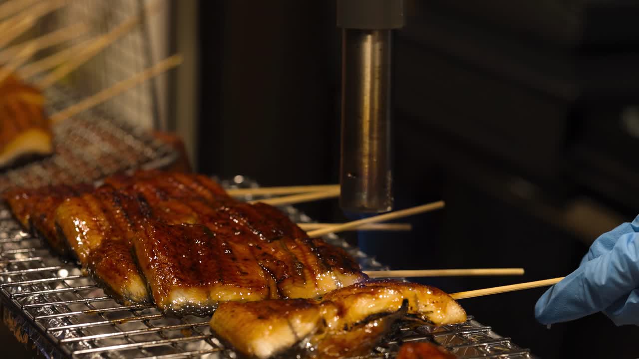 A chef uses a blowtorch to caramelize skewered meat, achieving a glossy finish on a metal grill.