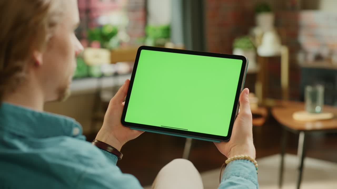 joven sosteniendo una tableta con pantalla verde. hombre relajándose en casa, viendo videos y leyendo publicaciones en las redes sociales en un dispositivo móvil. imágenes cercanas sobre el hombro.