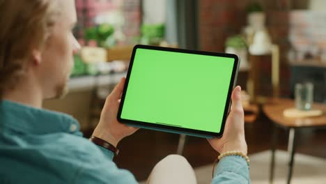 young man holding tablet computer with green screen mock up display. male relaxing at home, watching videos and reading social media posts on mobile device. close up over the shoulder footage.
