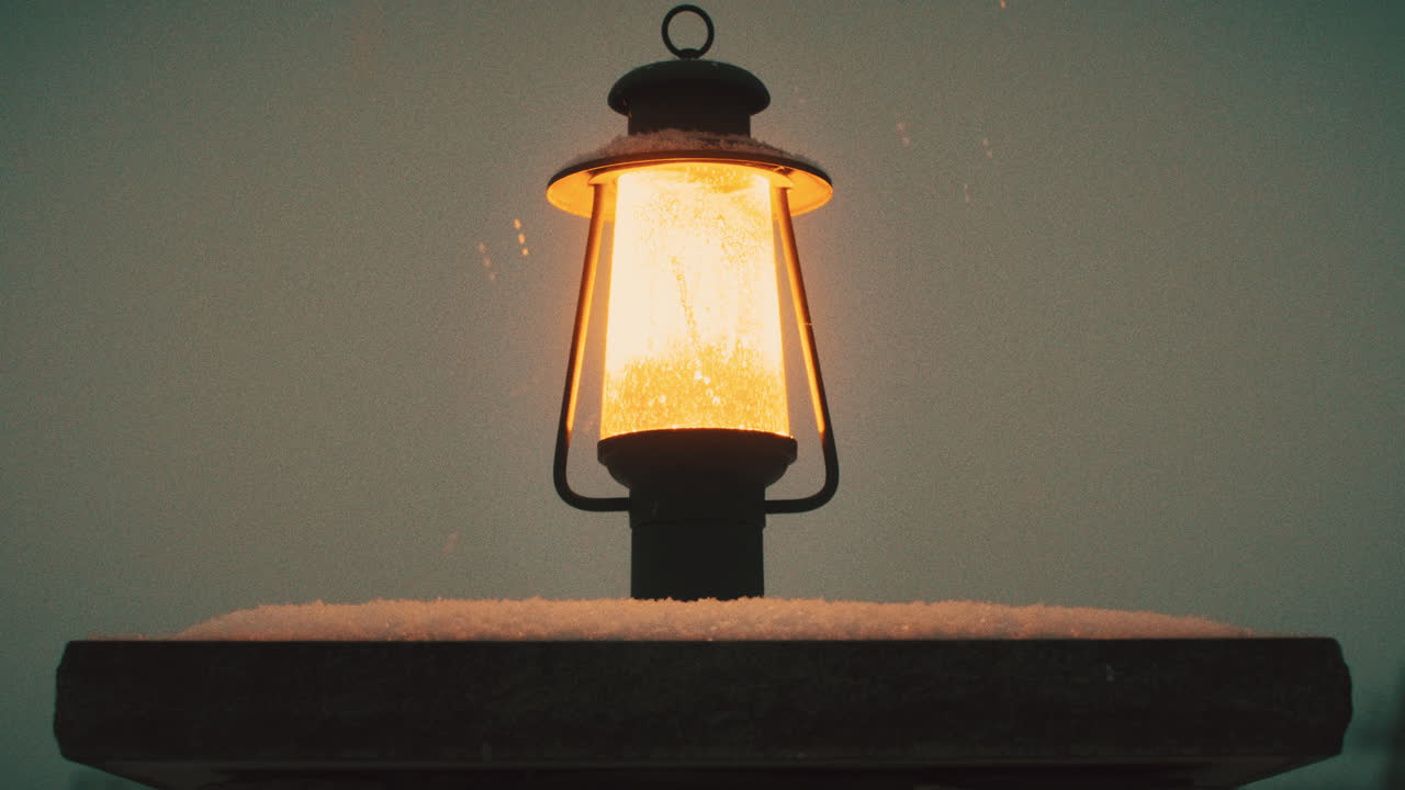 Inquadratura statica di un lampione posizionato su una colonna di cemento che illumina con luce gialla mentre nevica al crepuscolo
