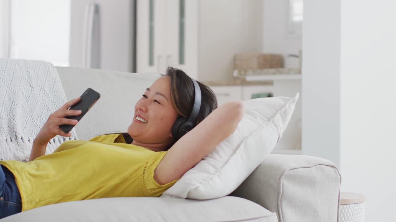 소파에 누워 헤드폰을 착용 한 행복한 아시아 여성, 집에서 스마트 폰으로 휴식을 취하고