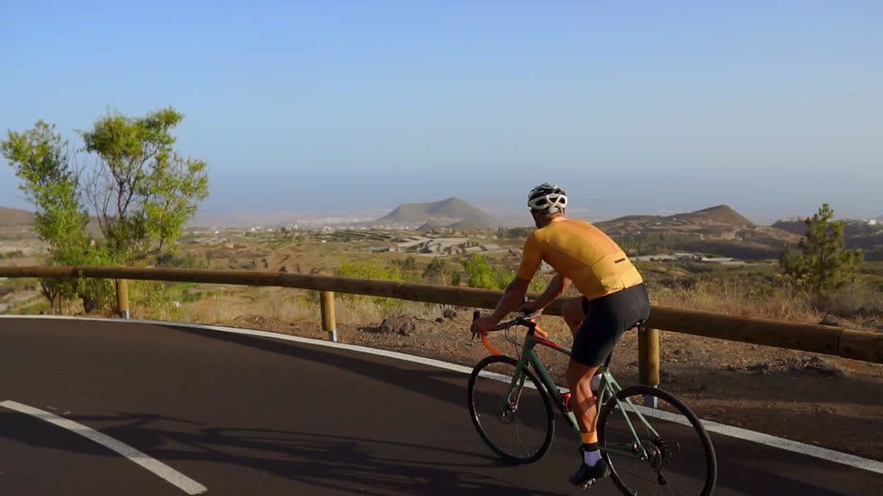 Tracking video shot of a male cyclist climbing up a mountain road. Man doing cycling training on a hilly highway road on a sunny day.