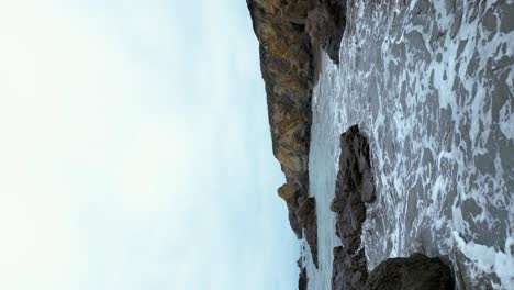 Vertical-shot-of-waves-crashing-on-shore-along-pacific-coastline,-San-Mateo-County-California-Highway-1