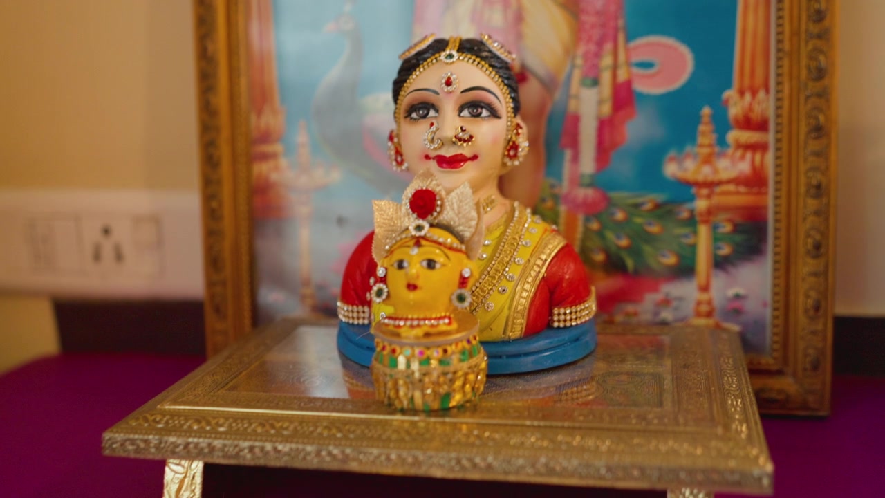 Ídolo de boneca de casamento colorida do sul da Índia posicionado em frente a um pôster emoldurado de divindade hindu em uma plataforma decorada