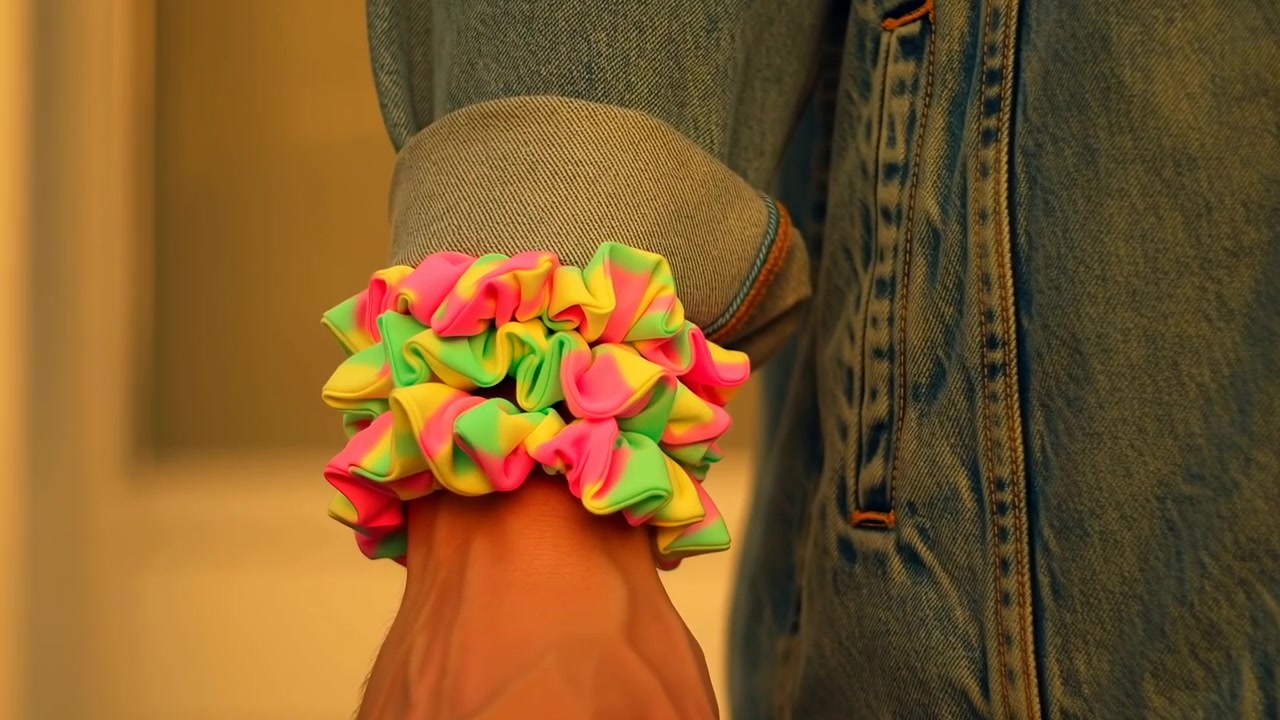 Muñeca con scrunchie rosa neón, amarillo y verde que descansa bajo el puño enrollado de una chaqueta vaquera, bajo una cálida luz interior, revelando volantes comprimidos, costuras, textura de la piel y detalles de estilo casual