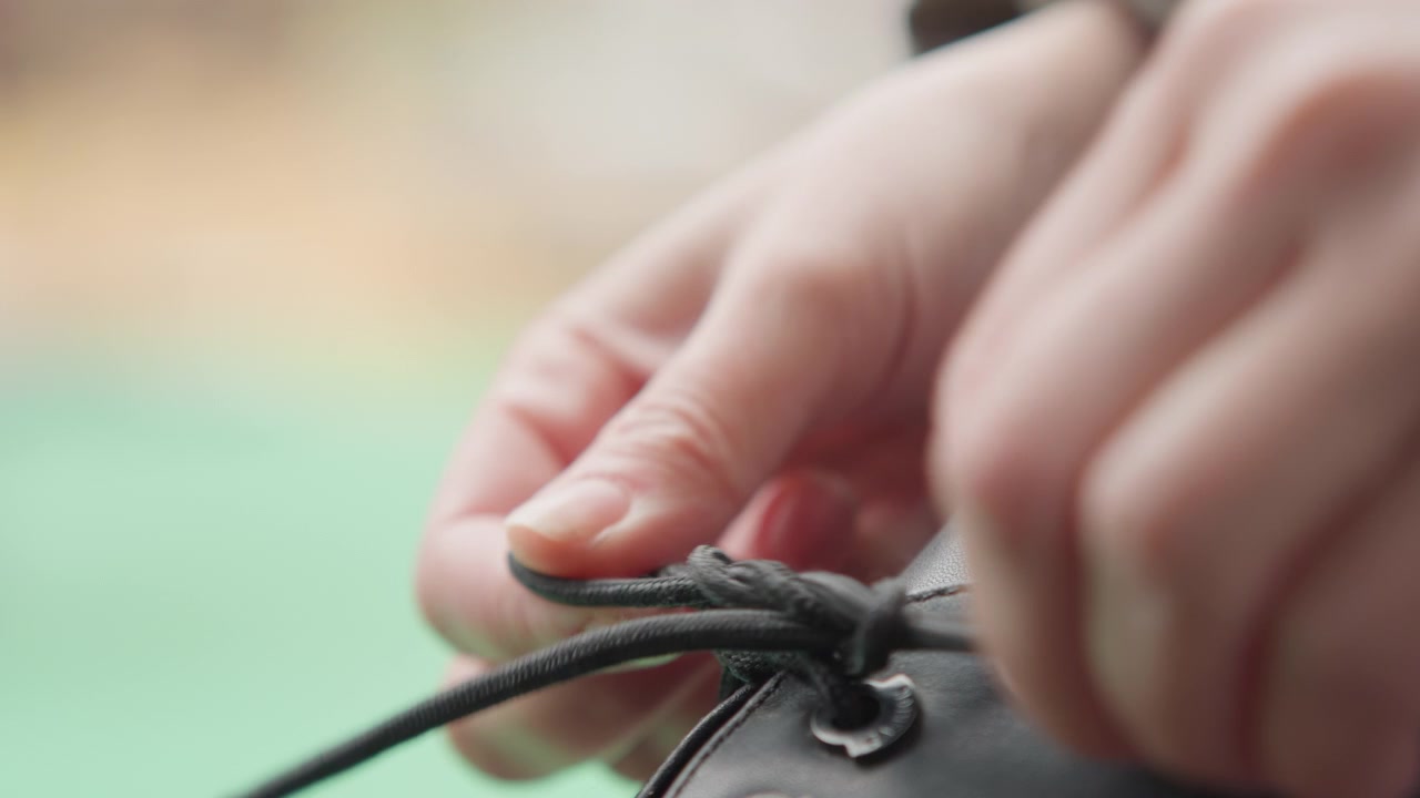 Closeup hands finishing lace bow, intimate tutorial mood with soft background, careful finger movements forming neat knot and loop, emphasis on secure finish, texture of leather and aglet visible