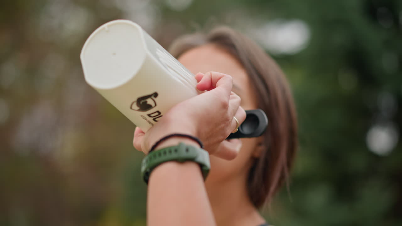 Close-up of woman drinking water from white bottle with blurred background, promoting hydration, outdoor fitness, wellness, and active lifestyle, while enjoying a refreshing drink