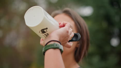 close-up of woman drinking water from white bottle with blurred background, promoting hydration, outdoor fitness, wellness, and active lifestyle, while enjoying a refreshing drink