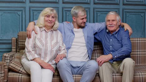 Familia-De-Abuelos-Mayores-Con-Un-Hijo-Adulto-Abrazándose-Y-Divirtiéndose-Mirando-La-Cámara-Uniéndose-En-Casa