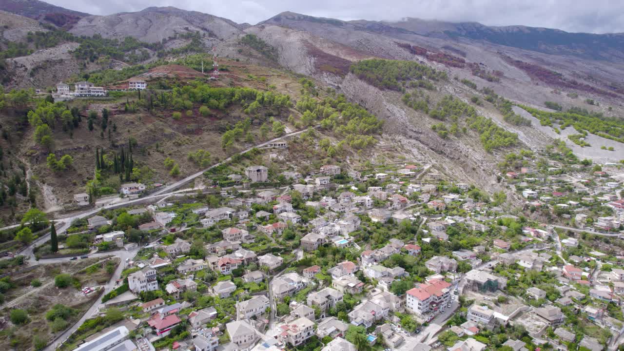 Un filmato realizzato con un drone mostra una piccola città residenziale con case sparse e vegetazione alla base di una catena montuosa rocciosa sotto un cielo nuvoloso, con un movimento fluido della camera.