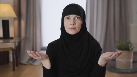 portrait of young muslim lady sitting with raised hands, looking at camera and talking. young woman in traditional black hijab praying at home. religion, traditional culture.