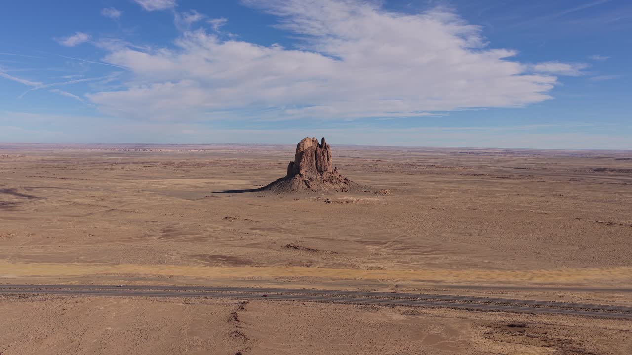 Un volo di drone si muove verso un'unica butte desertica e un viaggio in auto su autostrada con un'auto che attraversa un vasto paesaggio arido con formazioni geologiche aspre sotto un cielo azzurro brillante.