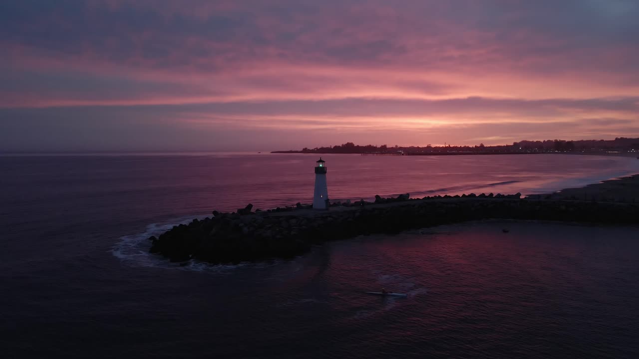 A drone shot circling the Walton Lighthouse in Santa Cruz California in front of a stunning sunset with a kayaker leaving the Pacific Ocean and entering the small crafts harbor