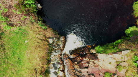 Aerial-top-down-shot-starting-close-to-a-small-waterfall-then-rising-to-reveal-stream-and-grass-around-it,-bright-summers-day