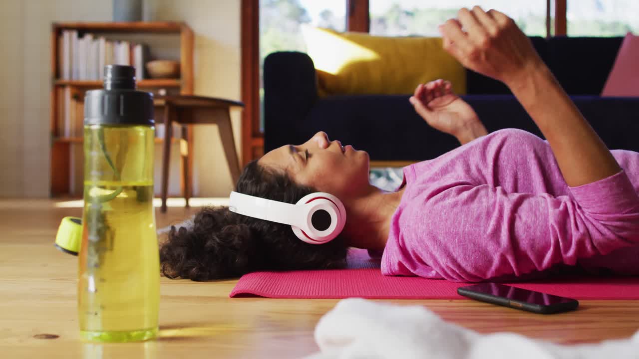mujer de raza mixta con auriculares practicando yoga y meditando mientras está acostada en la alfombra de yoga en casa