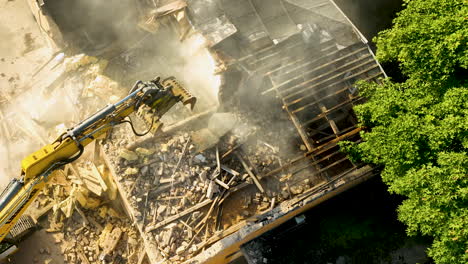 high-angle shot of an excavator claw dismantling a residential building's roof, scattering debris across the site