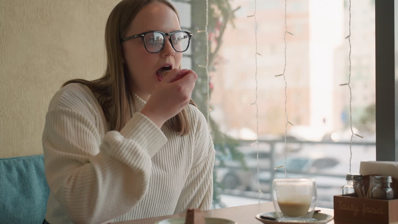 close up of young woman in white sweater slicing chocolate cake in cafe with latte on wooden table, decorative lights hanging on window with soft blur of snowy city street and cars in background