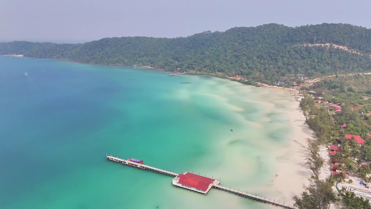Drone pulls away over turquoise waters and white sandy beach of Kaoh Rong Sanloem, Cambodia, revealing the long pier, dense coastal forest, and the island's gently sloping tropical hills